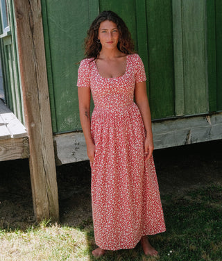 Woman in a red floral dress standing in front of a green wooden building.