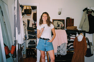 A woman stands in a doorway wearing a white t-shirt and denim shorts, holding a small black object. The background shows a cluttered closet and dressing area with clothes, bags, and shelves.