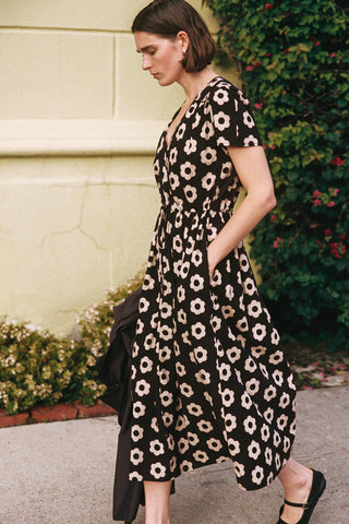 Woman wearing a black floral dress standing outdoors with greenery in the background