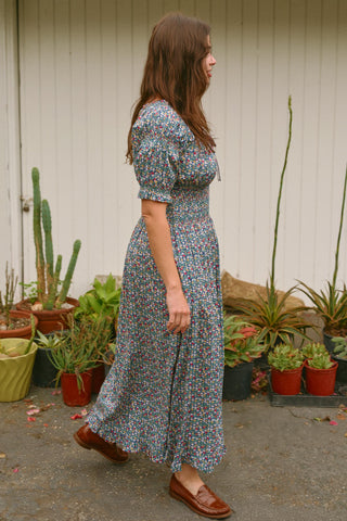 Woman in a floral dress standing among potted plants against a white wall.
