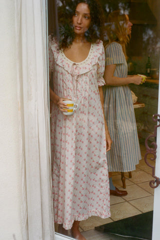 Woman in a floral dress holding a cup, standing in a doorway with another person in the background.