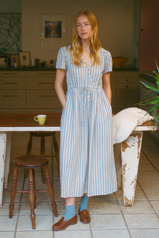 Woman in a striped dress standing in a kitchen with wooden furniture and a plant.