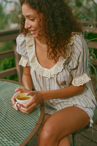 Woman sitting outdoors holding a cup, wearing a striped shirt with lace details.