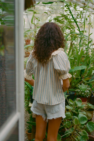 Person in a stripe blouse and denim shorts standing in a greenhouse surrounded by plants