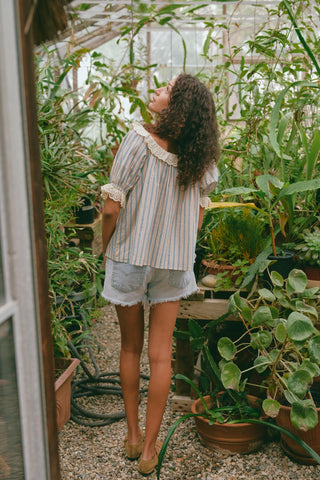 Person in a stripe blouse and denim shorts standing in a greenhouse surrounded by plants