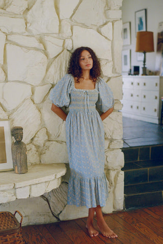 Woman in a blue dress standing in a room with stone walls and wooden floors.