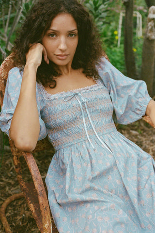 Woman in a light blue dress sitting outdoors with greenery in the background