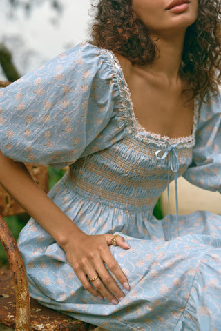 Woman wearing a light blue dress with puff sleeves and floral patterns, sitting outdoors.