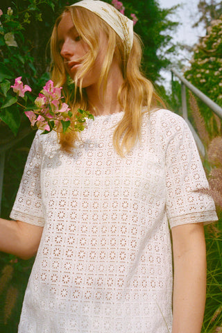Woman wearing a white lace tunic standing outdoors with greenery and flowers in the background