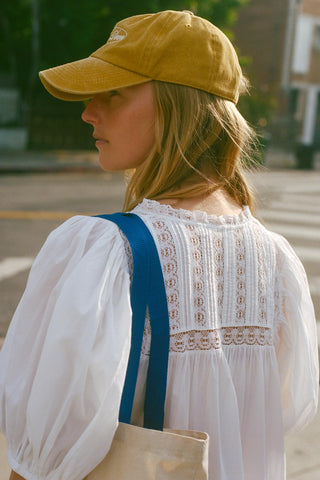 Person wearing a yellow cap and white blouse with blue straps, standing outdoors.