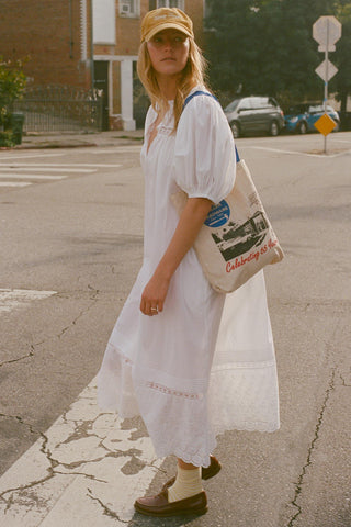 Woman in a white dress and yellow cap holding a tote bag on a street.