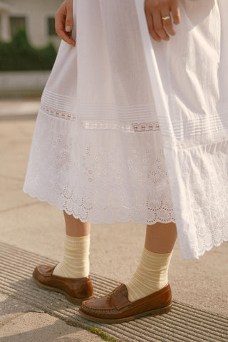 Person wearing a white dress with lace details, brown shoes, and striped socks on a sidewalk.