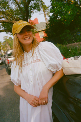 Woman in a white dress and yellow cap standing next to a car with trees and buildings in the background.