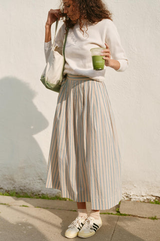 Woman in a white sweater and striped skirt holding a green cup against a white wall.