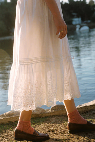 Person wearing a white lace dress and brown shoes by a body of water.