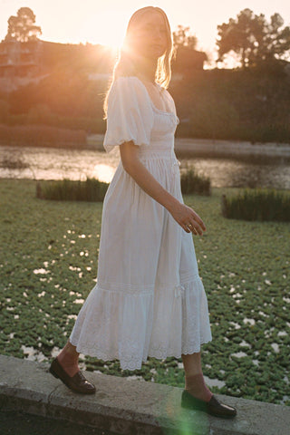 Woman in a white dress standing by a pond with sunlight filtering through.