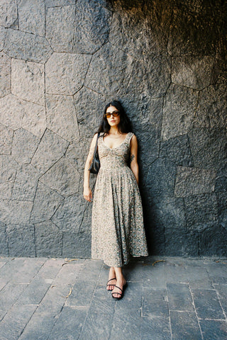 Woman in a green patterned dress standing against a stone wall wuth sunglasses on