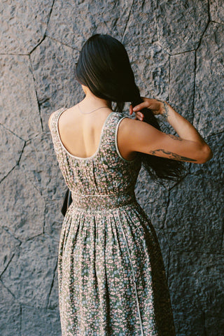 Back shot of a Woman in a green patterned dress standing against a textured stone wall.