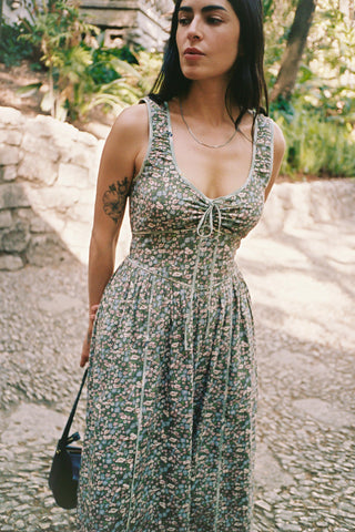 Woman wearing a green floral dress standing outdoors with stone wall and plants in the background