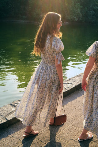 Two women in floral dresses standing by a body of water with sunlight filtering through trees.