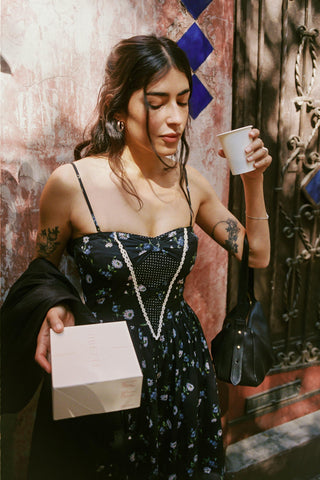Woman in a floral dress holding a cup and a box against a textured wall.