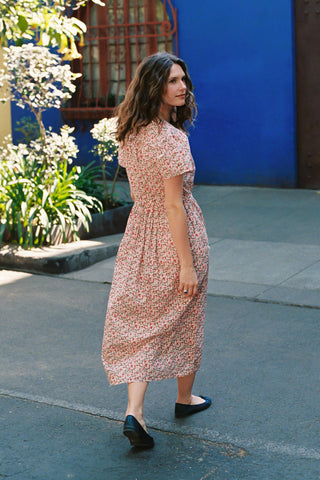 Woman in a floral dress standing on a sidewalk with a blue wall and plants in the background