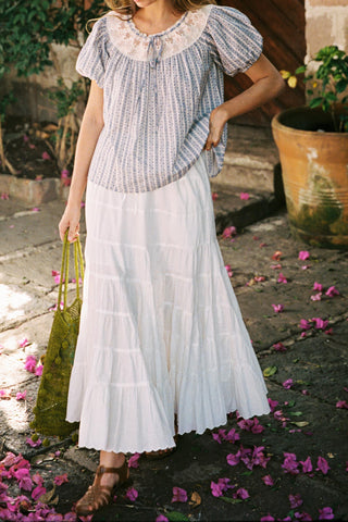 Woman in a striped blouse and white skirt standing on a stone path with flowers around.