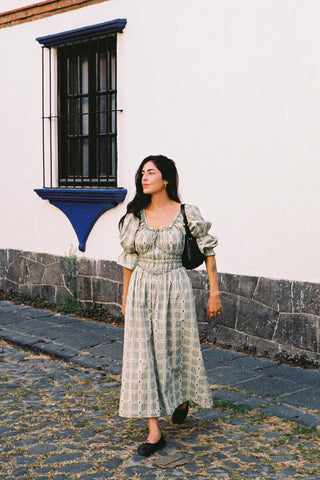 Woman in a green floral and plaid dress standing on a cobblestone street with a white wall and blue window frame in the background.