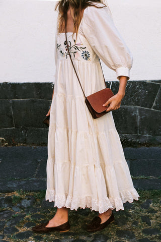 Woman wearing a white dress with floral embroidery, holding a brown leather handbag against a stone wall.