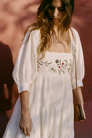 Woman wearing a white dress with floral embroidery against a pinkish-brown background