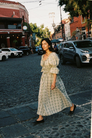 Woman in a long green floral and plaid dress  walking on a street with cars and buildings in the background