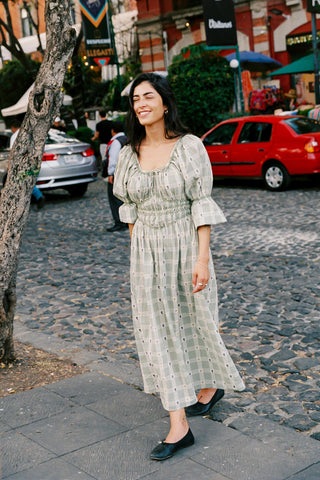 Woman in a green floral and plaid dress standing on a street with a red car and buildings in the background