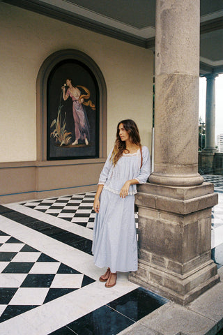 Woman in a light blue outfit standing next to a column in an elegant interior setting.