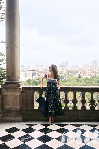 Woman in a polka dot dress standing on a balcony with a cityscape view.