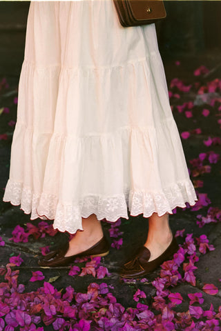 White lace skirt and black flats on a dark surface with purple flowers