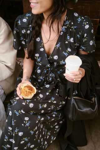 Woman in a dark floral dress holding a pastry and a coffee cup.