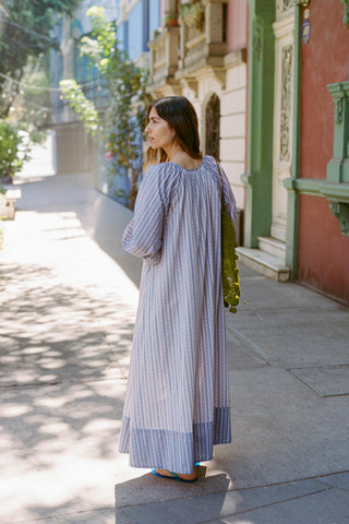 Woman in a long, light blue striped dress walking on a sunlit street.