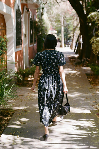 Woman in a black floral dress walking down a sunlit street with trees and buildings.