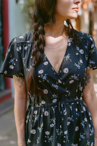 Woman wearing a black floral dress with tattoos on her arms, standing outdoors.