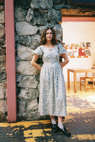 Woman in a floral dress standing against a stone wall indoors.