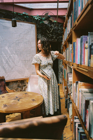 Woman in a floral dress standing in a cozy bookstore with bookshelves and tables.
