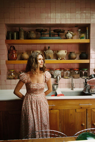 Woman in a floral dress standing in a kitchen with pink tiled walls and wooden cabinets.