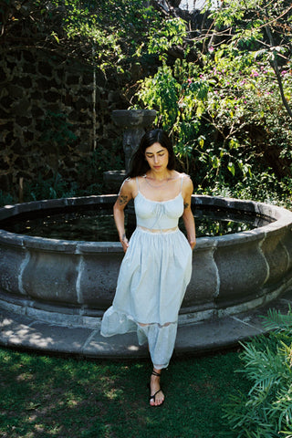 Woman in a light blue dress standing by a stone fountain in a garden.