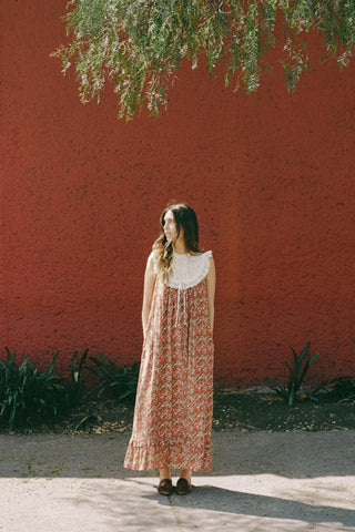Woman in a floral dress standing against a red wall with greenery.