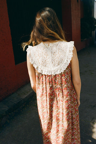 Woman wearing a floral dress with a white lace bib, standing against a red wall.