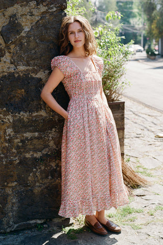 Woman in a floral dress standing against a stone wall on a street.