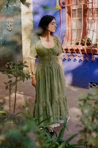 Woman in a green dress standing in a garden with plants and a colorful structure in the background.