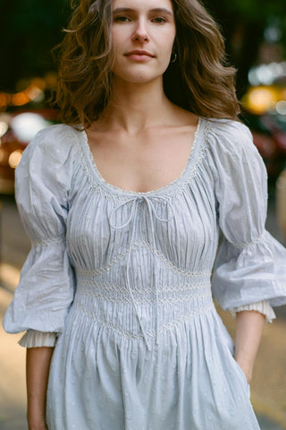 Woman wearing a light blue dress with smocked bodice and puffed sleeves against a blurred background