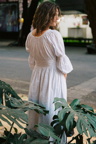 Woman in a light blue dress standing among green plants on a street.