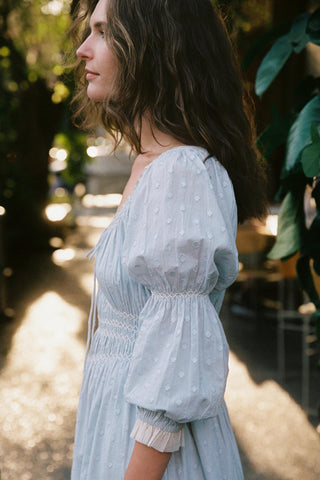 Woman in a light blue dress standing outdoors with greenery in the background
